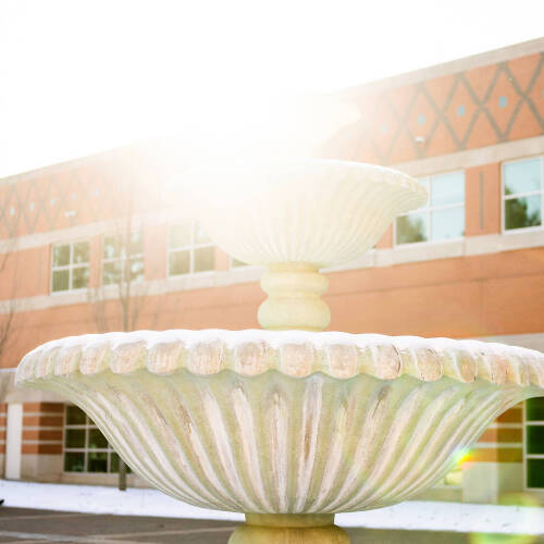 A snow-dusted fountain stands in front of a brick building with people walking by on a sunny winter day.